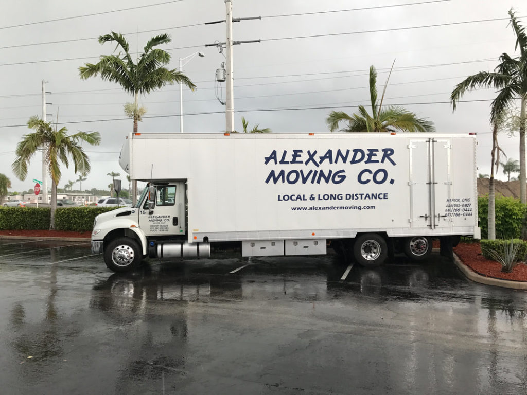 Alexander Moving Co. truck parked on wet lot. Palm trees. Overcast sky. Local & long distance service advertised.